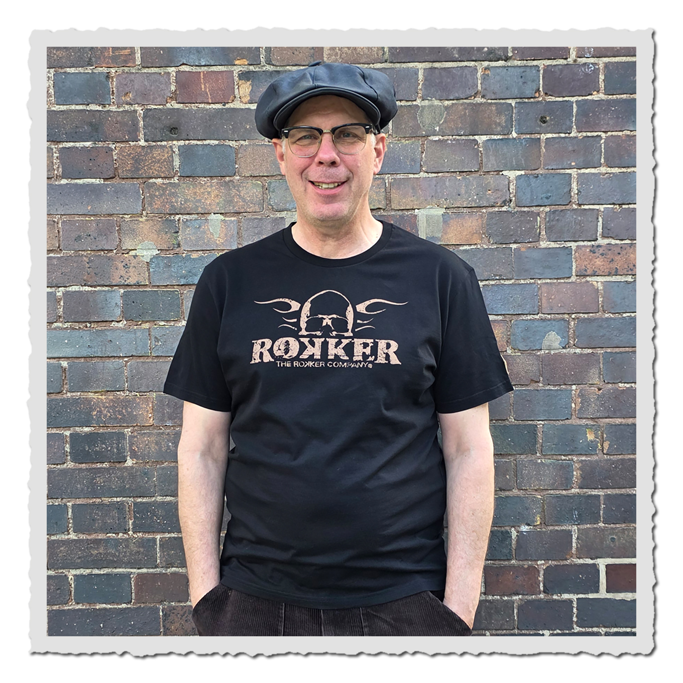 Mann von vorn in schwarzem T-Shirt mit hellem Brustprint, dunkler Kappe, vor mehrfarbiger Backsteinwand.